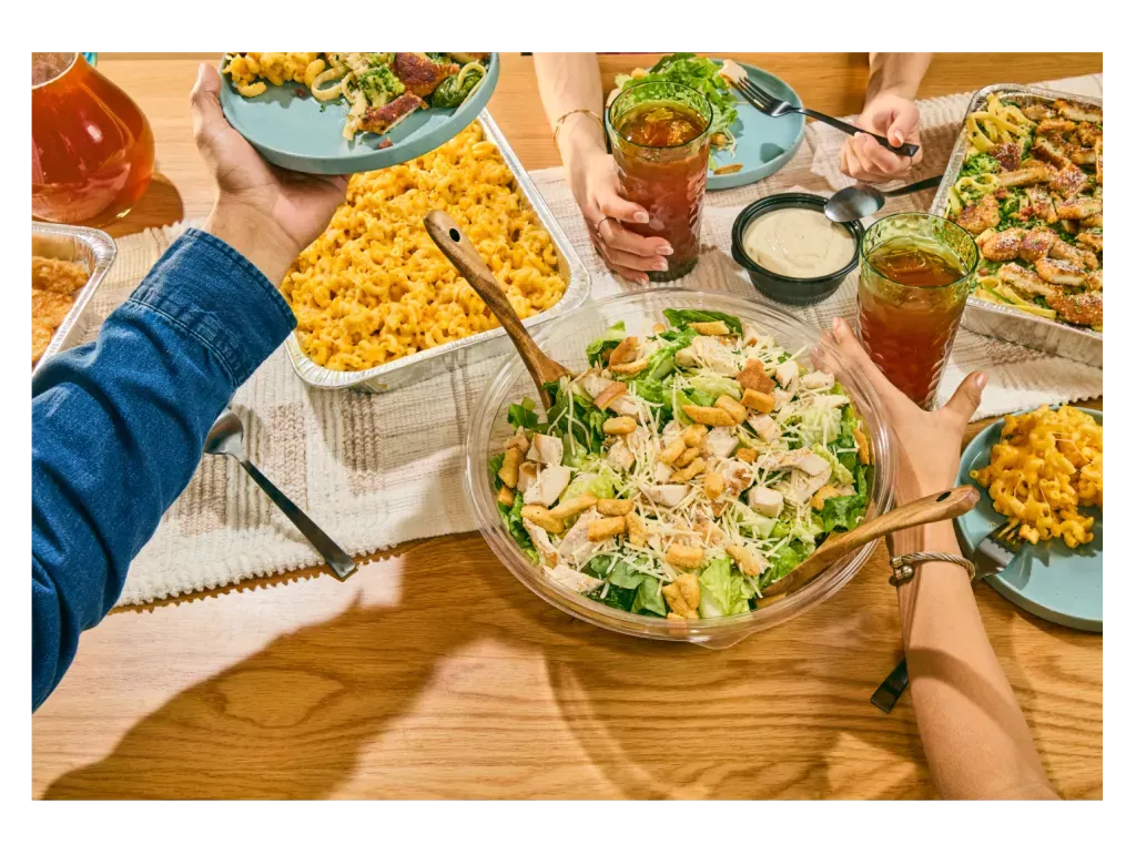 Catering dishes from Noodles and Company, including a pan of Creamy Cheddar Mac and Cheese, a Caesar Chicken Salad, and drinks