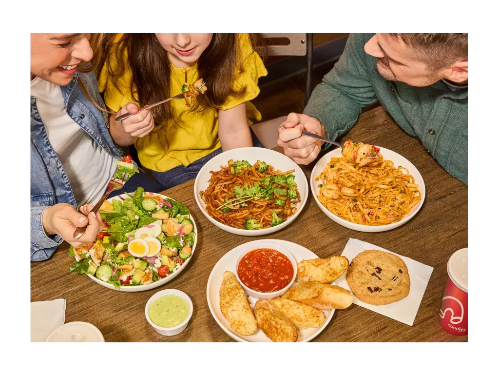3 people eating Noodles &amp; Company food, including a Green Goddess Cobb Salad, Japanese Pan Noodles, Pulled Pork BBQ Mac and Cheese, and Cheesy Garlic Bread.
