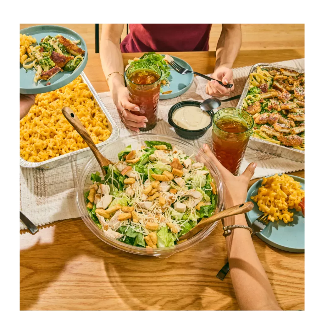 Noodles &amp; Company Catering dishes including a mac and cheese pan, alfredo pan, and large Caesar salad, with individual servings shown on blue plates