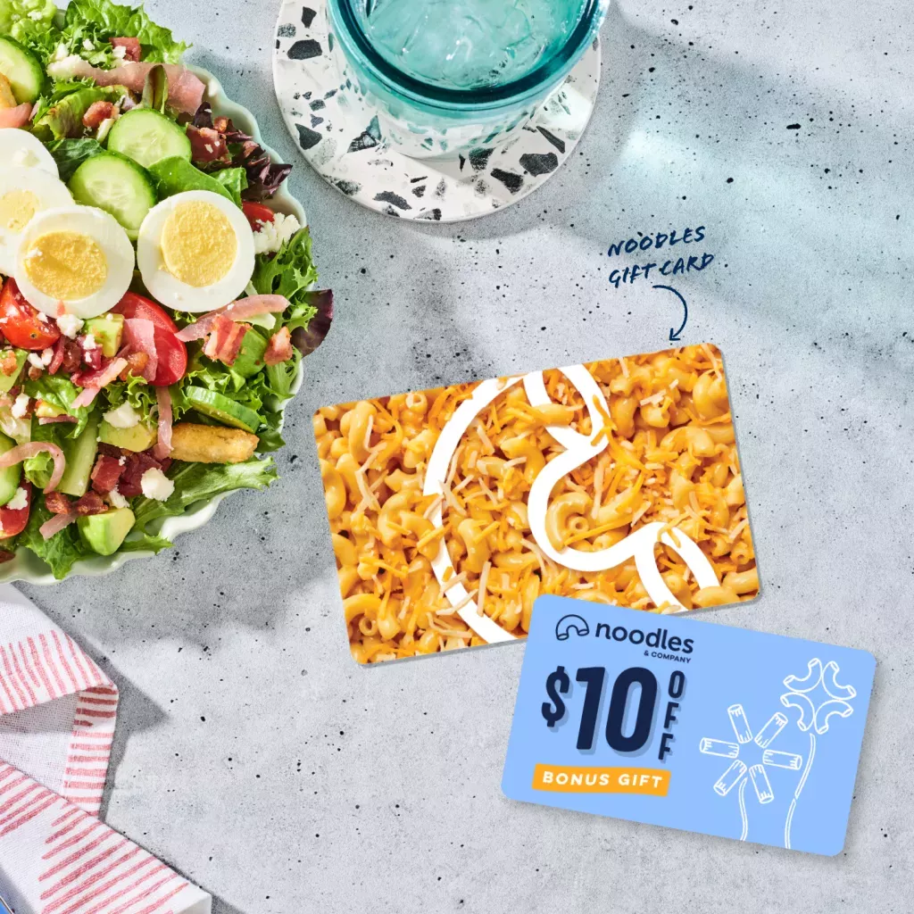 Noodles Gift Card with a $10 Bonus Card. Shown with a Green Goddess Cobb Salad, a napkin, and a glass of water in a blue cup.
