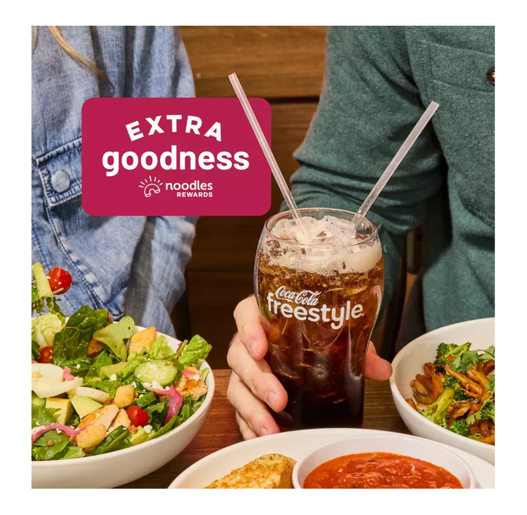 Extra Goodness from Noodles Rewards. Two people enjoying Noodles &amp; Company dishes with a Coca Cola Freestyle fountain drink with ice. Shown with a Green Goddess Salad, Japanese Pan Noodles, and a side of Cheesy Garlic Bread.