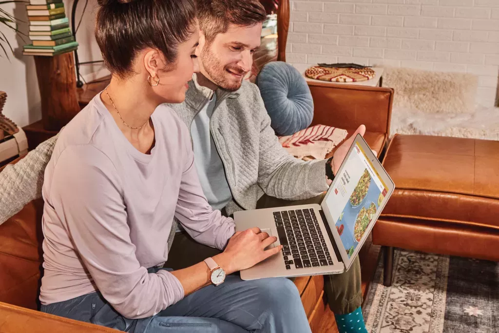 Noodles &amp; Company customers sitting on a couch in a living room browsing the Noodles &amp; Company website on a laptop.
