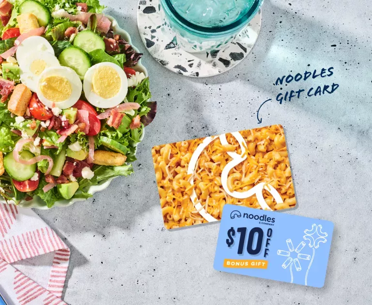 Noodles Gift Card with a $10 Bonus Card. Shown with a Green Goddess Cobb Salad, a napkin, and a glass of water in a blue cup.