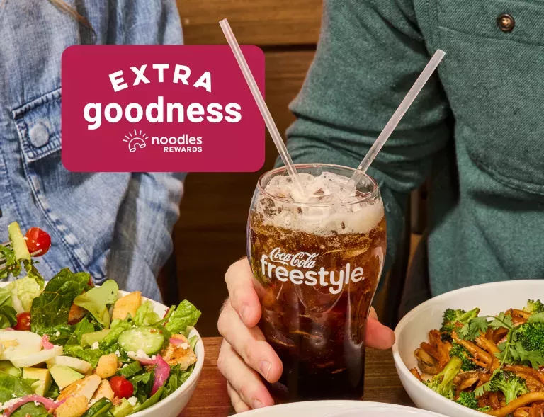 Extra Goodness from Noodles Rewards. Two people enjoying Noodles &amp; Company dishes with a Coca Cola Freestyle fountain drink with ice. Shown with a Green Goddess Salad, Japanese Pan Noodles, and a side of Cheesy Garlic Bread.