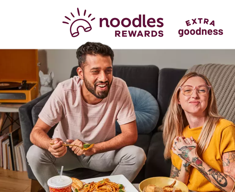 Noodles Rewards Extra Goodness. Two people enjoying Noodles and Company noodle dishes and appetizers on a couch.