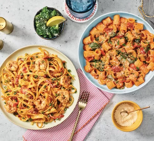 Entrees of Lemon Garlic Shrimp Scampi and Rigatoni Rosa on tabletop with a side of Lemon Parmesan Broccoli and grated parmesan.