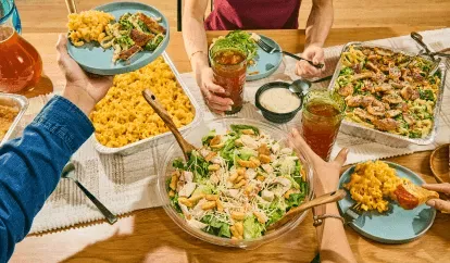 Noodles Catering on a wooden table. hands holding a beverage cup and a another handing a plate. Dishes featured include Caesar Salad, Creamy Cheddar Mac &amp; Cheese, and Crispy Chicken Bacon Alfredo