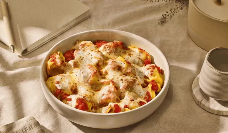 Baked Cheese Tortelloni on a cream-colored tablecloth next to a notebook, pen, a cup and candle.