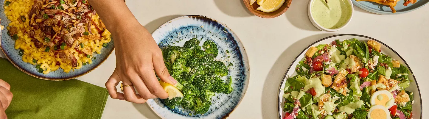 Tabletop image of a hand squeezing a lemon on parmesan broccoli next to a green goddess cobb salad and pulled pork BBQ mac and cheese.