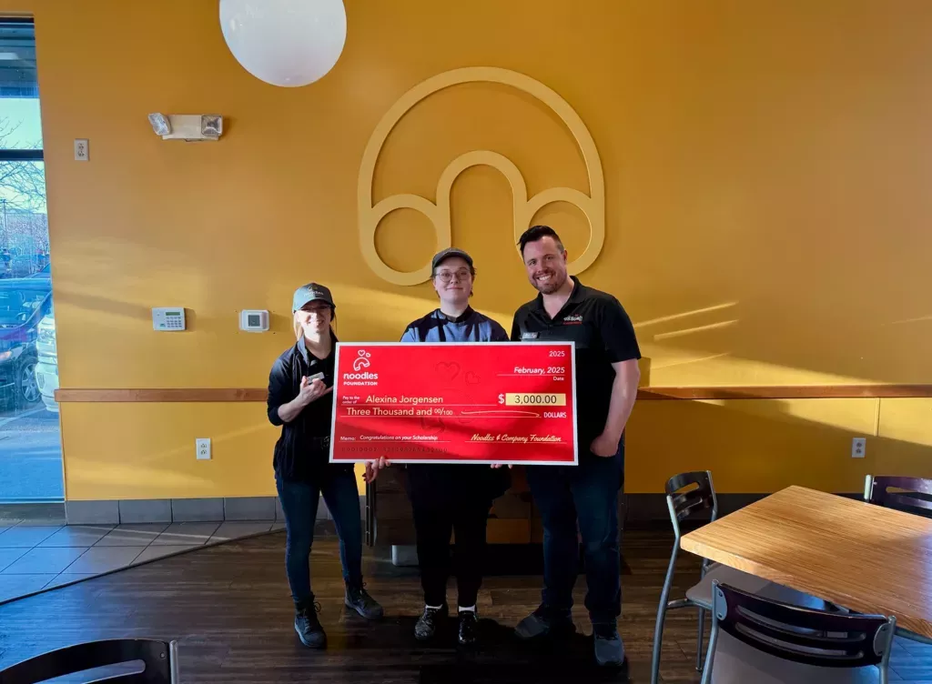 Noodles and Company 2025 Scholarship Award Winner holding a large check surrounded by friends, family and fellow employees.