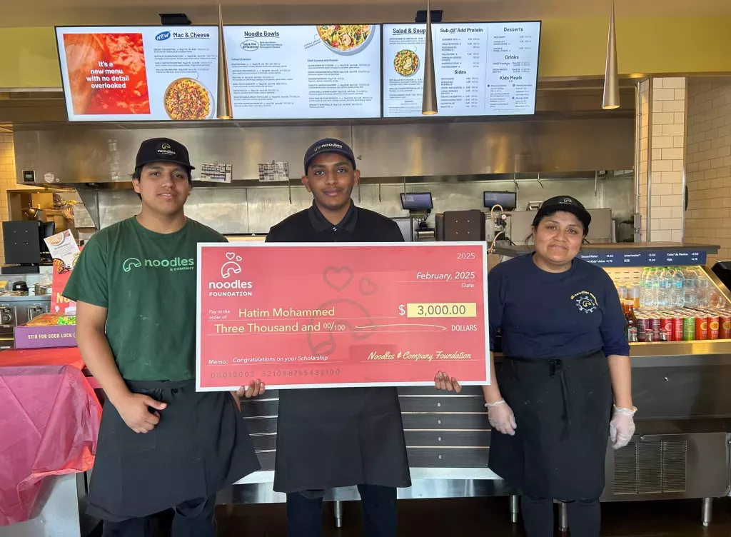 Noodles and Company 2025 Scholarship Award Winner holding a large check surrounded by friends, family and fellow employees.