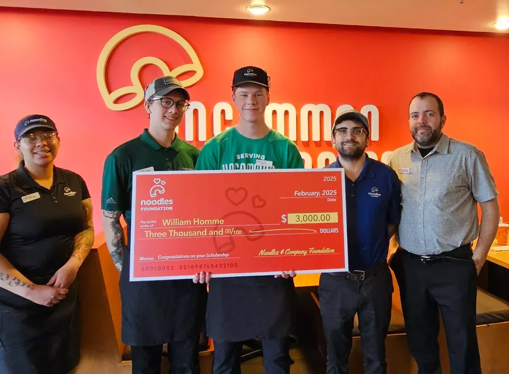 Noodles and Company 2025 Scholarship Award Winner holding a large check surrounded by friends, family and fellow employees.